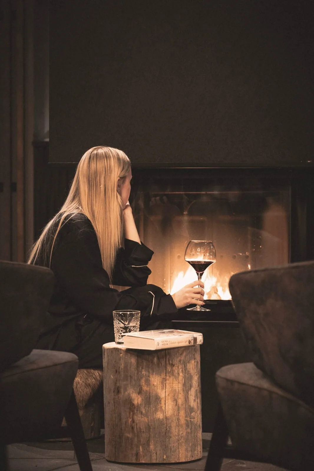 A guest sitting by the open fire, holding up a glass of red wine, with a book and a tumbler on a birch-round side table.
