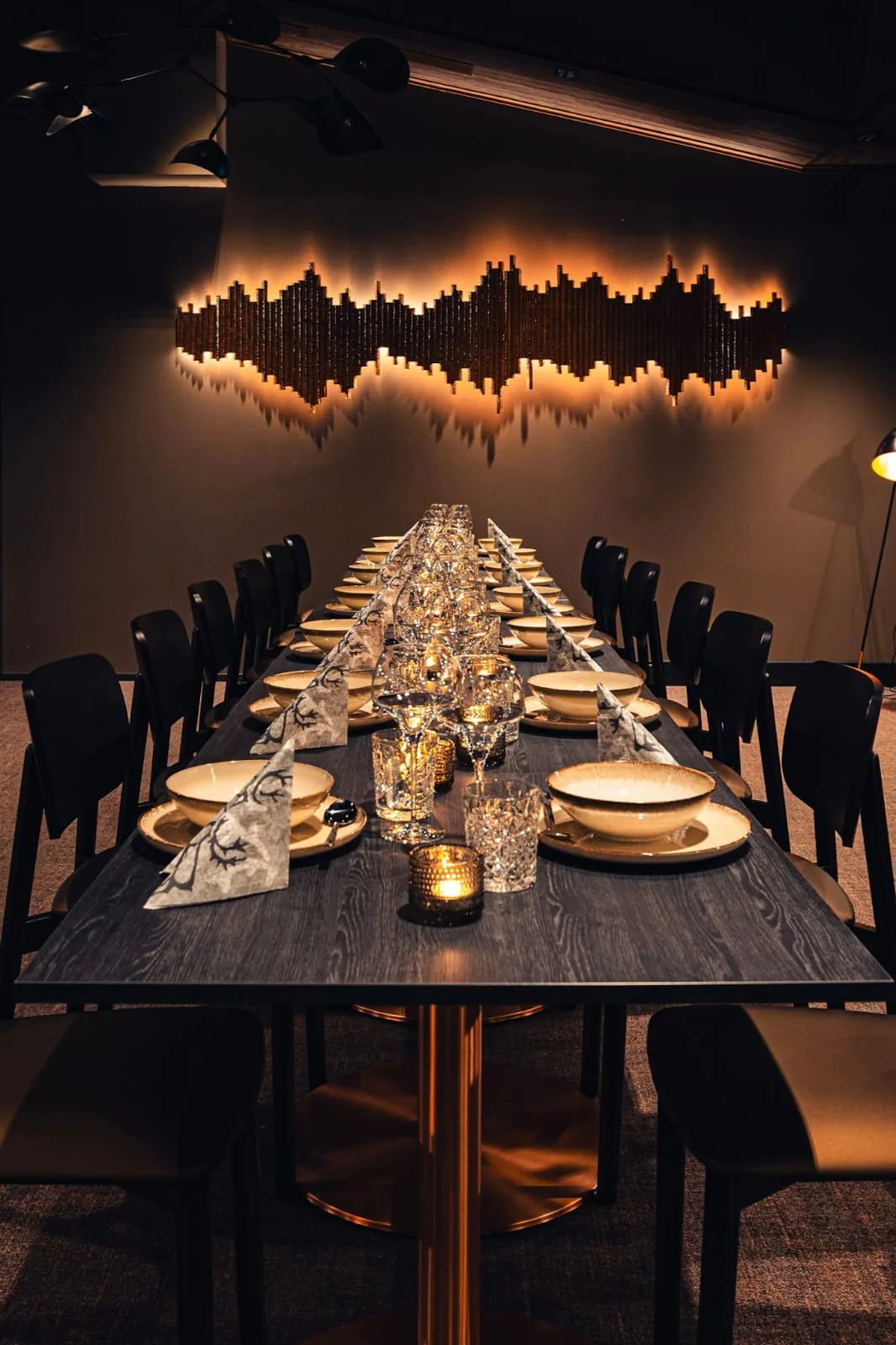 The long dining table laid for dinner: candles down the centre, glassware and gold-rimmed plates, dark chairs along both sides, and a backlit relief on the wall behind.