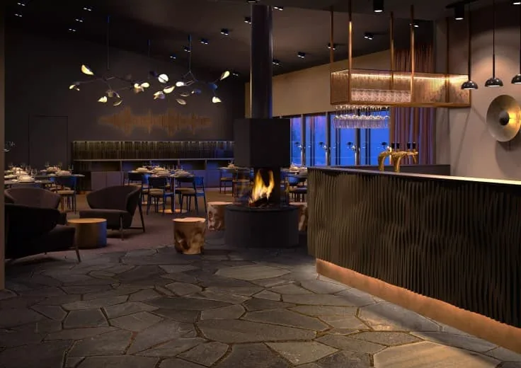 The bar: a fluted dark counter with brass taps and a copper-framed glass rack, a central open fire, log stools, dining tables beyond, and an irregular grey flagstone floor.