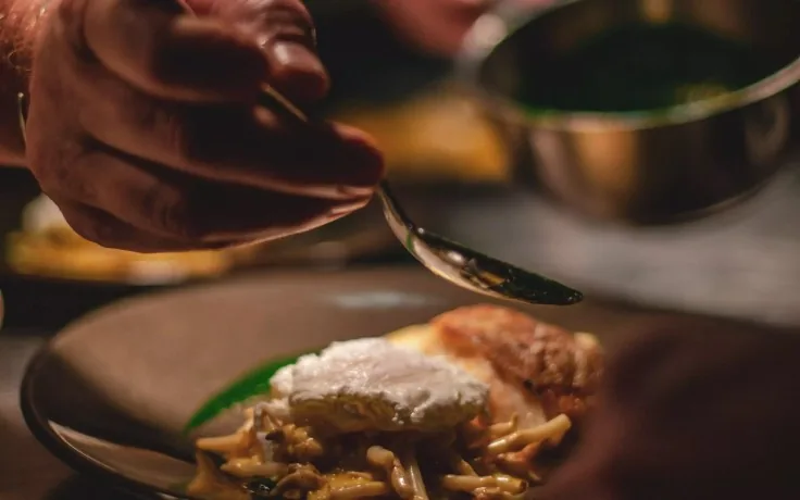 A cook's hand spooning dark herb oil over a plated dish: a poached egg and a crisp-edged fillet on braised enoki mushrooms, with a steel bowl of green oil behind.