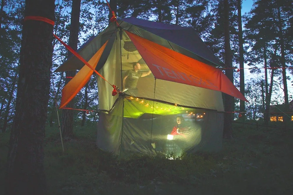 A person sitting inside a lit tree tent at dusk, the tent hung clear of the ground with small lights strung along it.