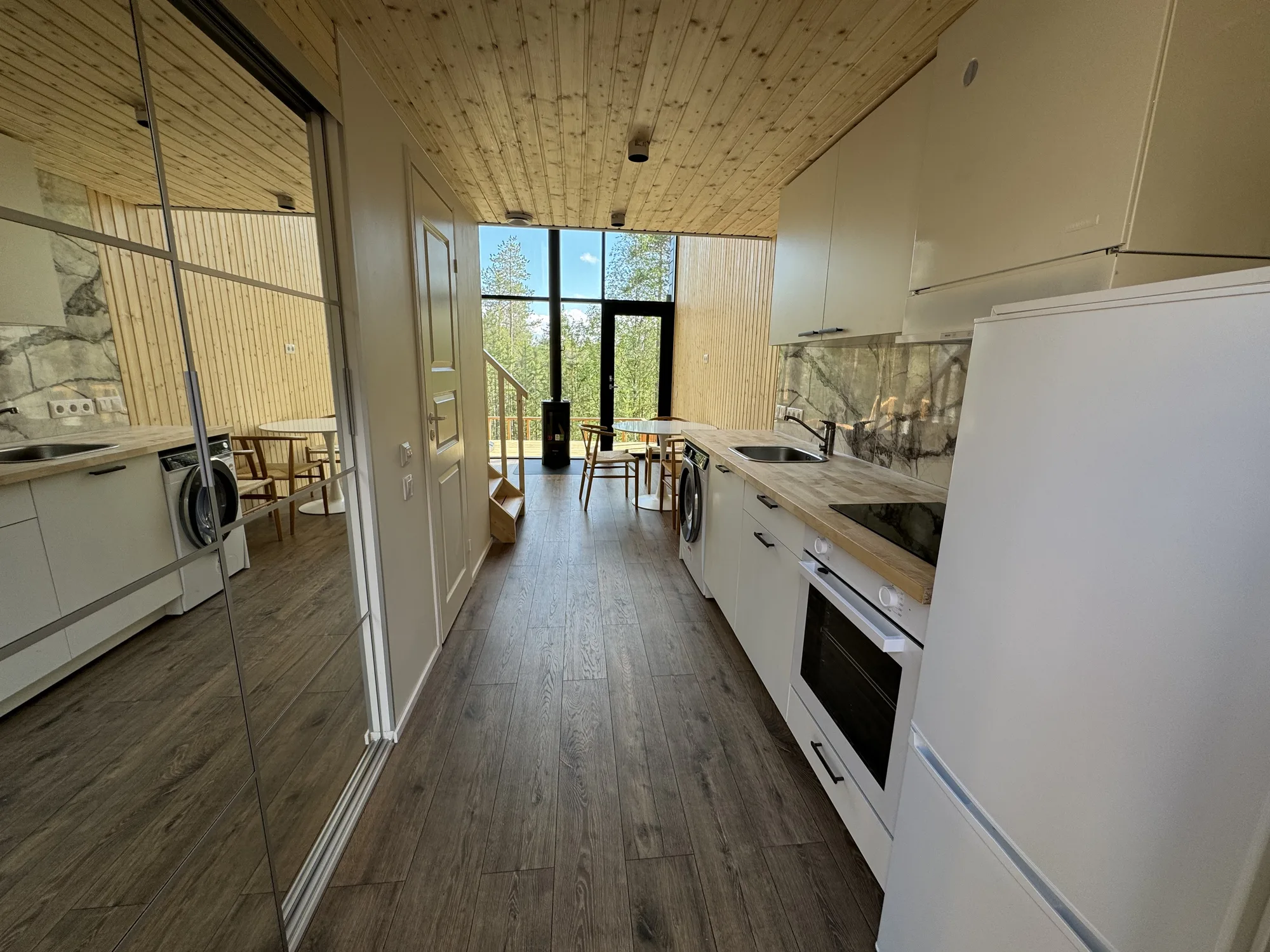 Owl's Nest open-plan interior: a white kitchen along one wall, a dining table, a black fireplace and tall glazing onto green forest.