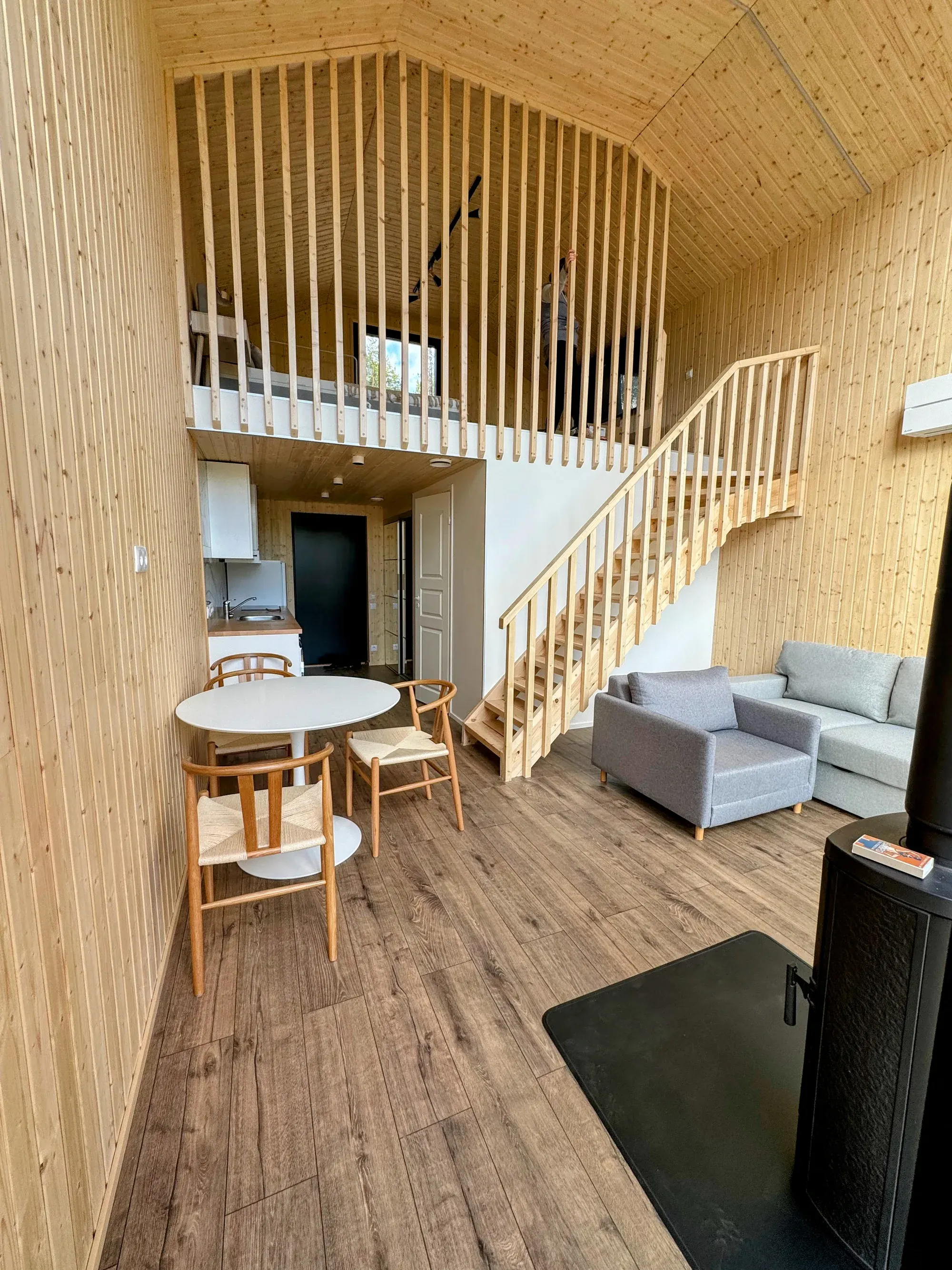 Owl's Nest double-height living room with a timber loft, slatted balustrade, staircase, black wood stove and dining table beside tall forest-facing windows.