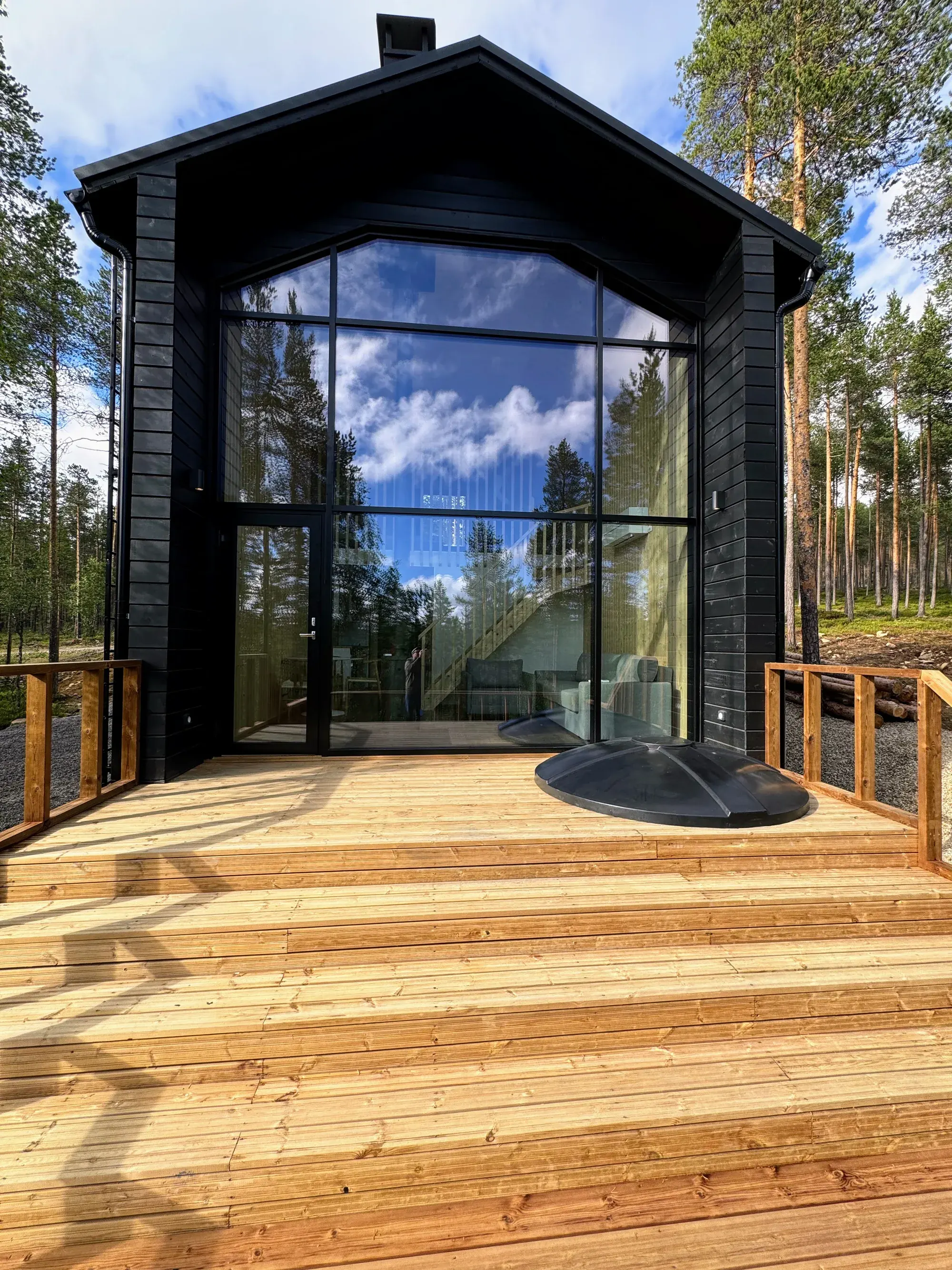 Owl's Nest in summer: its black timber gable, full-height glass wall, timber deck and sunken hot tub framed by pine forest.