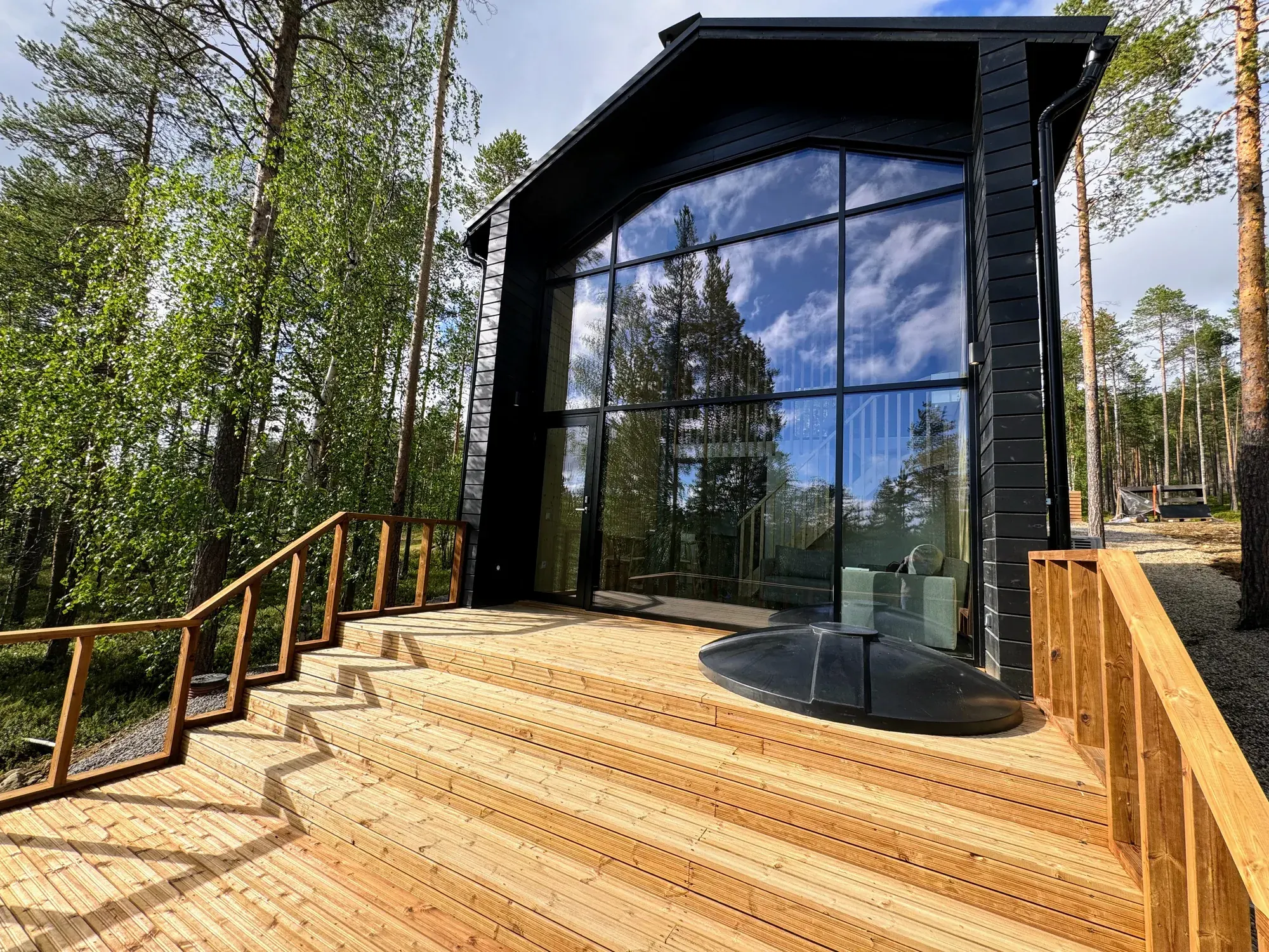 Owl's Nest in summer: a black timber chalet with a full-height glass gable, a broad timber terrace and a round outdoor hot tub among pine and birch trees.