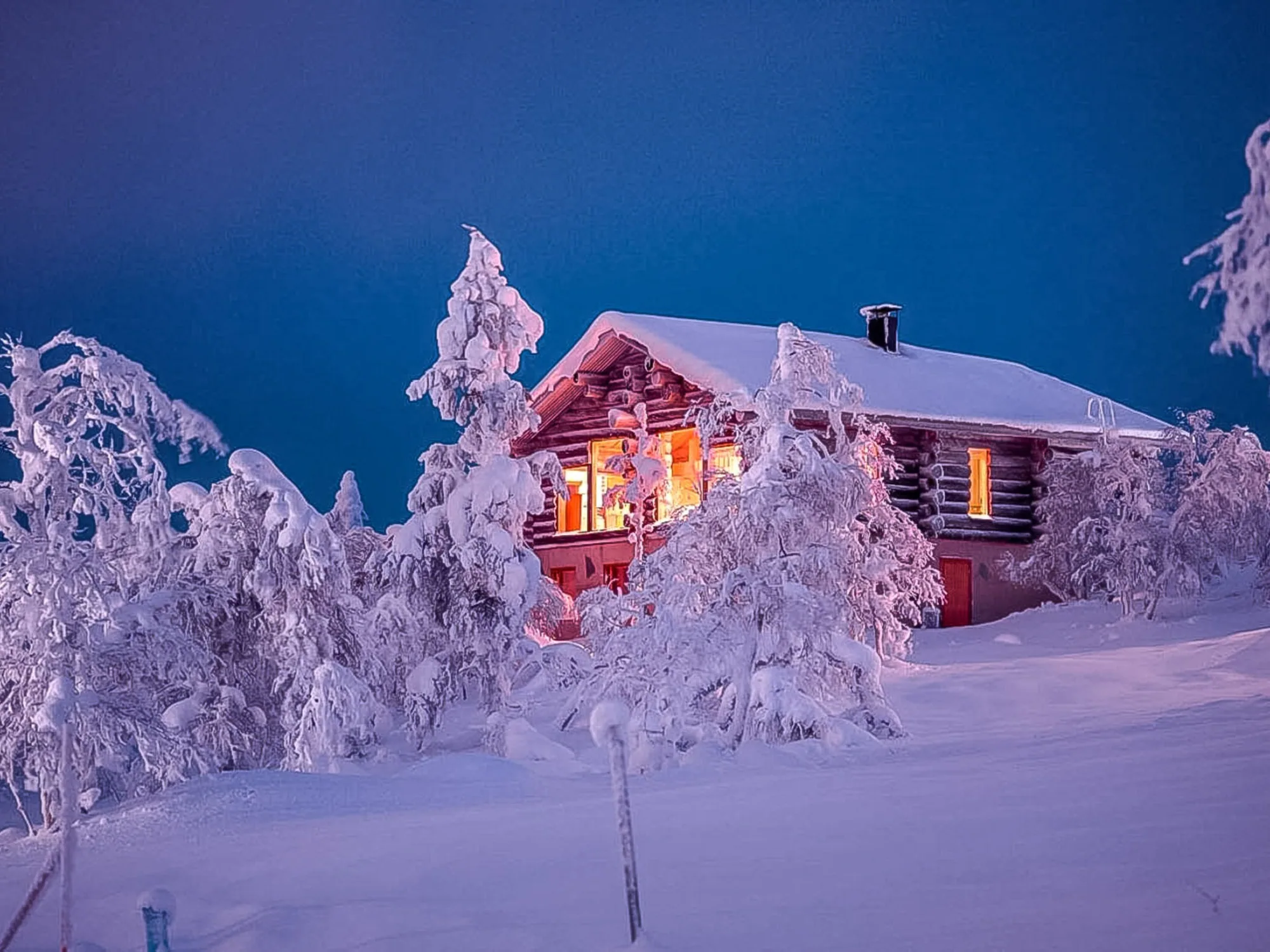 A log chalet at blue hour, its windows lit gold, standing in deep snow among heavily frosted trees.