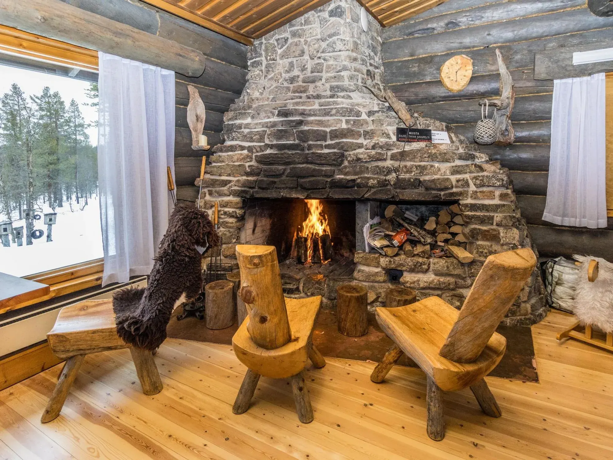 A stone fireplace with a live fire, hand-hewn log chairs and a bench on a pine floor, snow visible through the window beside it.